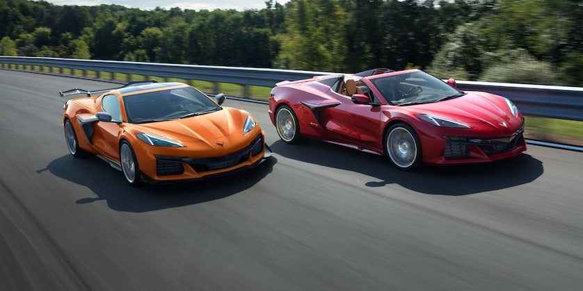 Orange car with roof and Red Chevrolet Corvette without roof running on road near jungle